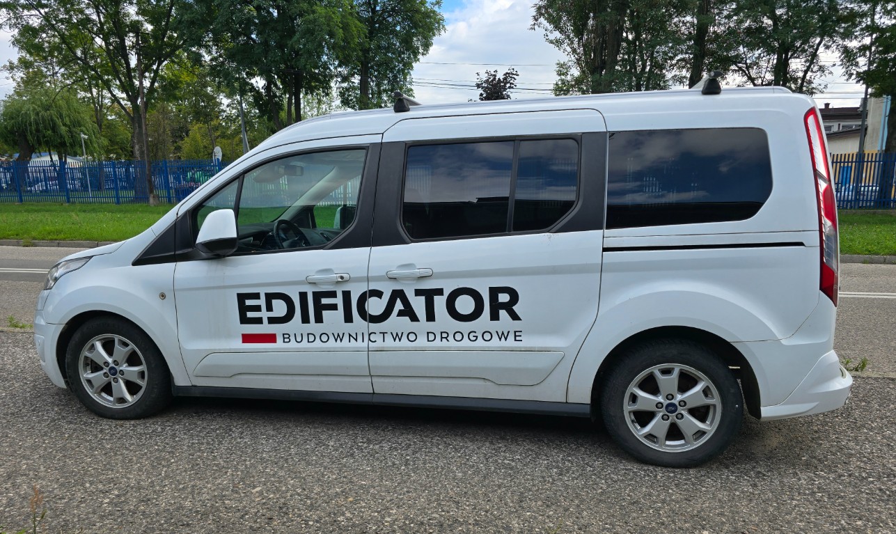 Biały furgon Ford z napisem „EDIFICATOR BUDOWNICTWO DROGOWE” na boku, zaparkowany na asfalcie. W tle zielone drzewa, niebieskie ogrodzenie i chmurne niebo. Pojazd ma czarne szyby i srebrne felgi.